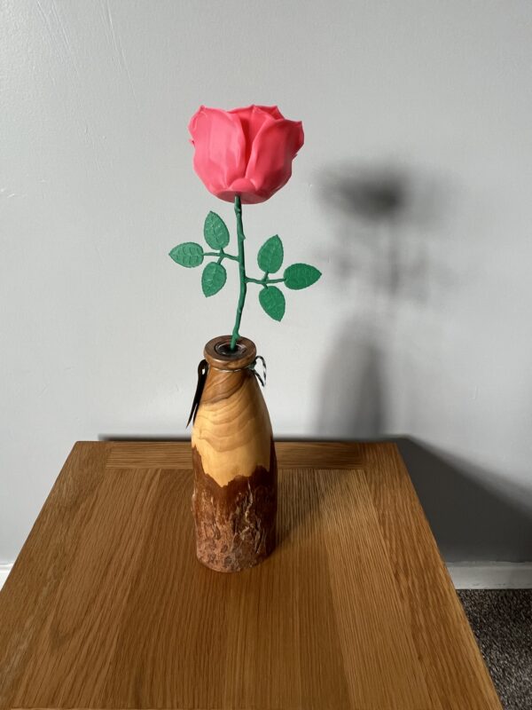 A vibrant pink 3D-printed rose with green leaves, elegantly displayed in a handcrafted wooden vase, illustrating the blend of traditional craftsmanship and innovative 3D printing